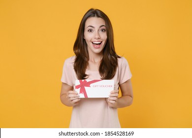 Excited surprised young brunette woman 20s wearing pastel pink casual t-shirt posing holding in hands gift certificate looking camera isolated on bright yellow color wall background studio portrait