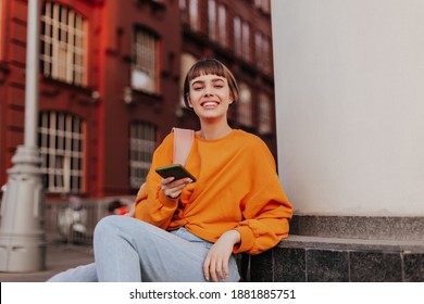 Excited teen girl in orange sweatshirt leans on building and holds smartphone. Joyful woman in stylish hipster outfit smiles outside. - Powered by Shutterstock