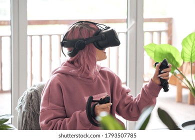 Excited teen girl pink hair wears vr headset goggles holds controllers plays vr video game futuristic immersive simulator explores virtual reality 3d 360 cyber gaming experience sits in chair at home.