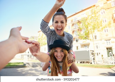 Excited Teenager On Back Of Happy Friend In Hat Laughing As They Reach For A Hand From First Person Perspective As Photographer