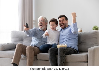 Excited Three Generation Men Family Fans Hold Remote Control Watch Tv Sport Game Sit On Sofa Together, Happy Grandson Young Father And Old Grandfather Celebrate Goal Television Team Victory At Home