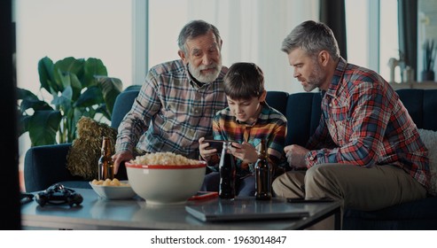 Excited Three Generation Men Family Grandson Young Father And Old Grandfather Fans Hold Soccer Ball Remote Control Watch Tv Sport Game Celebrate Goal Television Team Victory In Tv Match Sit On Sofa