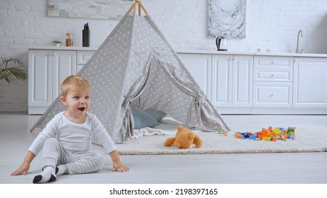 Excited Toddler Boy Sitting Near Baby Wigwam And Toys On Carpet