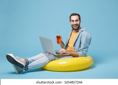 Excited Traveler Tourist Man In Yellow Clothes Isolated On Blue Background. Passenger Traveling Abroad On Weekend. Air Flight Journey Concept. Sit In Inflatable Ring, Work On Laptop, Hold Beer Bottle