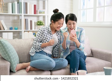 Excited Two Asian Women Friends Checking Good News In Smart Phone Sitting On Sofa In Living Room At Home. Beautiful Young Girls Relax On Couch With Yes Hand Gesture Looking On Cellphone Screen Indoor