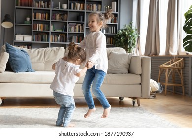 Excited two little girls children have fun jumping dancing in cozy living room on weekend. Happy small babies kids siblings hold hands engaged in funny activity at home. Entertainment concept.