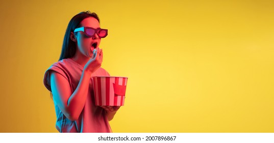 Excited. Watching cinema. Young pretty caucasian girl in 3d glasses with bucket of popcorn isolated on yellow studio background in neon light. Flyer. Copyspace for ad. - Shutterstock ID 2007896867