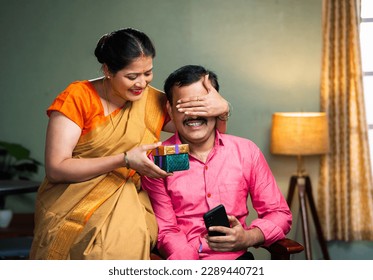 excited wife giving gift to husband by closing eyes while sitting on chair at home - concept of togetherness, happt long relationship and wedding anniversary. - Powered by Shutterstock