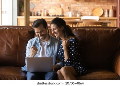 Excited Winners. Overjoyed Married Couple Sit On Large Sofa At Modern Cottage Interior Use Laptop App Laugh Win Lottery Prize. Amazed Young Spouses Raise Fists Yes Get Great Offer For Family Business