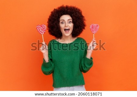 Excited woman with Afro hairstyle in green casual style sweater holding two candies with heart shape, looking at camera with happy positive expression. Indoor studio shot isolated on orange background