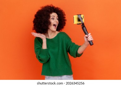 Excited Woman With Afro Hairstyle Wearing Green Casual Style Sweater Using Stedicam And Phone For Livestream, Waving Hand To Followers. Indoor Studio Shot Isolated On Orange Background.