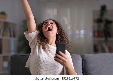 Excited Woman Celebrating Success Holding Mobile Phone Sitting On A Couch In The Night At Home