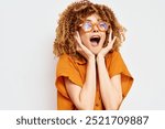 Excited woman with curly hair wearing orange glasses, expressing joy and surprise against a plain light background, showcasing happiness and vibrant emotions