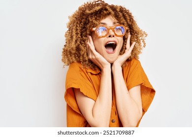 Excited woman with curly hair wearing orange glasses, expressing joy and surprise against a plain light background, showcasing happiness and vibrant emotions - Powered by Shutterstock