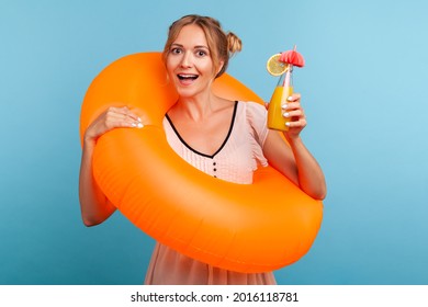 Excited Woman With Funny Hair Buns Relaxing At Summer Resort, Holding Orange Rubber Ring And Enjoying Fresh Fruit Cocktail, Laughing Happily At Vacation. Indoor Studio Shot Isolated On Blue Background