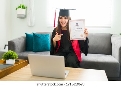 Excited Woman In Her 20s Showing Off A University Certificate To The Family During An Online Video Call. Pretty Female Graduate Wearing A Graduation Gown And Cap Sitting On The Living Room