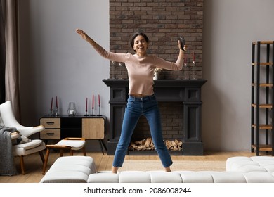 Excited Woman Holding Smartphone Jumping On Couch At Home, Overjoyed Young Female Having Fun Alone, Dancing Moving To Favorite Popular Music, Enjoying Leisure Time With Device In Living Room