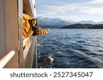 Excited woman leans out of a ferry, enjoying the fresh air and breathtaking snowy mountain scenery.