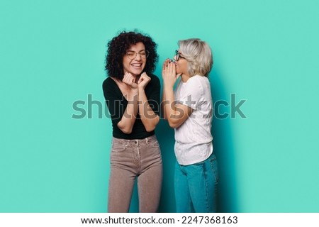Excited woman listening to friend whispering to her ear on the blue background. Fashion model. Attractive beautiful girl.