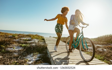 Excited Woman Riding Bike Down The Boardwalk With Her Friends Running. Two Young Female Friends Having A Great Time On Their Vacation.