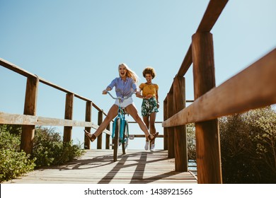 Excited Woman Riding Bike Down The Boardwalk With Her Friends Running Behind. Two Young Female Friends Having A Great Time On Their Vacation.