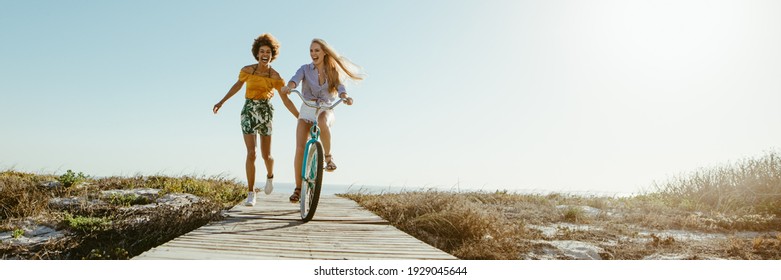 Excited woman riding bike down the boardwalk with her friends running along. Two female friends having a great time on their vacation. Panoramic shot with lots of copy space on background.