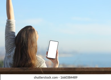 Excited Woman Showing Blank Smart Phone Screen Raising Arms Sitting On A Bench