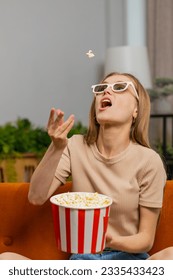 Una mujer emocionada sentada en el sofá arroja comiendo palomitas de maíz, trata de atrapar la boca. Niña con gafas 3D viendo interesantes series de televisión, juegos deportivos, películas, contenido de películas en medios sociales en línea en casa. Vertical