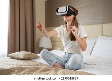 Excited Woman Sitting On King-size Bed Plays VR Games In Hotel Room. Happy Tourist Wearing Special Glasses For VR Games Explores Virtual World