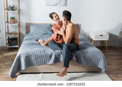 Excited Woman In T-shirt Near Shirtless Man Sitting On Bed In Pajama Pants