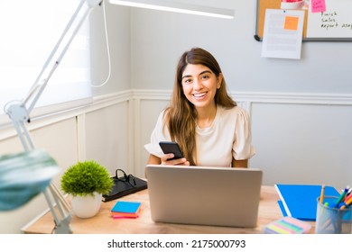 Excited woman or virtual assistant smiling and using her smartphone while doing her remote work at the office desk 