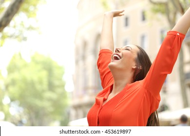 Excited woman wearing orange blouse raising arms outdoors in the street with buildings in the background - Shutterstock ID 453619897