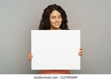 Excited young adult female hold blank placard looking down to empty copy space with pleasant smile. Beautiful millennial woman in pink outfit show while poster or banner isolated at studio background - Shutterstock ID 2127095849