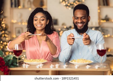 Excited Young African American Couple Sitting In Restaurant And Taking Pictures Of Food With Mobile Phones For Social Media, Foodie Bloggers Celebrating St Valentine's Day Together