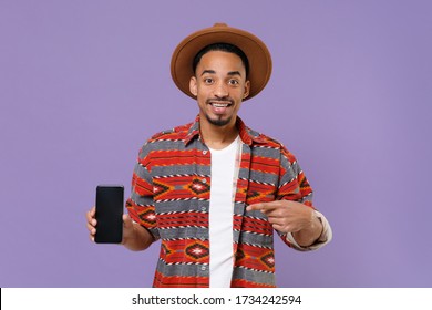 Excited Young African American Guy In Casual Colorful Shirt Hat Isolated On Violet Background. People Lifestyle Concept. Mock Up Copy Space. Point Index Finger On Mobile Phone With Blank Empty Screen