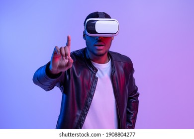 Excited Young African American Guy In VR Glasses And Leather Jacket Enjoying Virtual Reality Experience, Amazed Black Gamer Man Standing In Neon Lighting Over Purple Studio Background, Copy Space