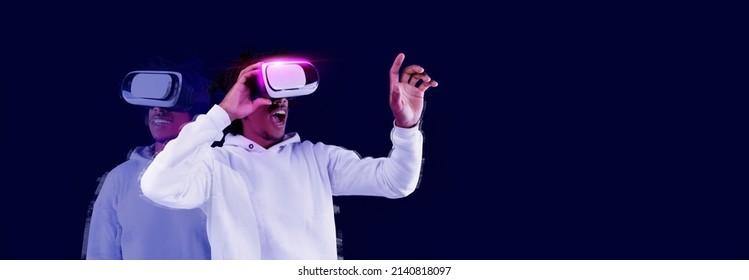 Excited young African American guy using VR glasses, touching something invisible at copy space, teeanger experiencing virtual reality isolated on blue neon studio background, panorama banner mockup