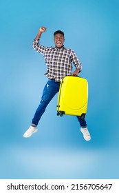 Excited Young African American Guy With Yellow Suitcase Jumping And Gesturing YES On Blue Studio Background, Full Length. Overjoyed Black Male Traveler With Luggage Flying Up In Air