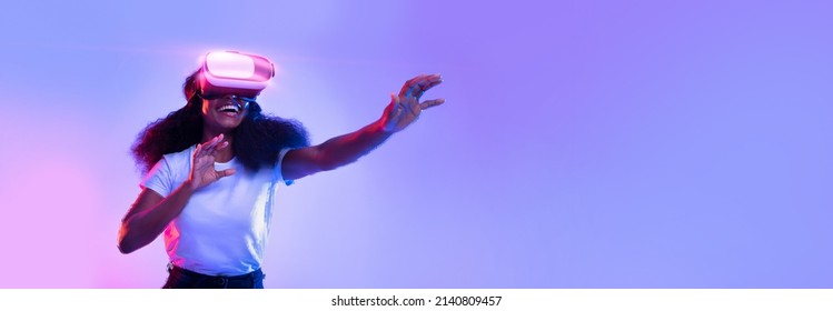 Excited young African American lady using VR glasses, touching something invisible at copy space, millennial female experiencing ar virtual reality adventure on neon studio background, panorama banner - Shutterstock ID 2140809457
