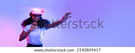 Excited young African American lady using VR glasses, touching something invisible at copy space, millennial female experiencing ar virtual reality adventure on neon studio background, panorama banner