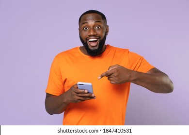 Excited Young African American Man 20s Wearing Basic Casual Orange T-shirt Pointing Index Finger On Mobile Cell Phone Typing Sms Message Isolated On Pastel Violet Colour Background, Studio Portrait