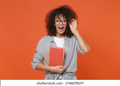 Excited young african american student girl in casual clothes, eyeglasses isolated on orange background studio portrait. People lifestyle concept. Mock up copy space. Hold books, keeping mouth open