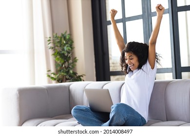 Excited Young African American Woman Yelling Yes, Sitting At Home On The Couch Looking At The Laptop Computer Screen, Celebrating Victory, Goal Achievement Raised Arms Up