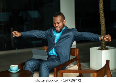 Excited Young African Businessman Sitting At A Table In The Lobby Of A Modern Office And Working On A Digital Tablet Arms Up