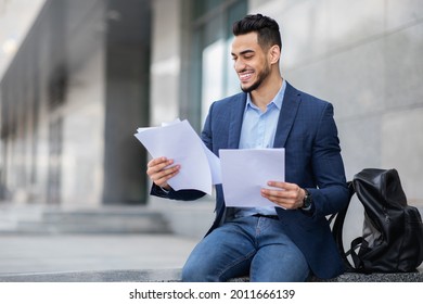 Excited young arab man in formal outfit sitting on the street next to business building, holding documents or CV, having backpack by him, getting ready for job interview, panorama with copy space - Powered by Shutterstock