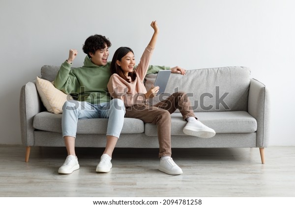 Excited\
young Asian couple sitting on couch with tablet pc, celebrating\
online win, great deal or business success at home, free space.\
Millennial spouses enjoying big sale in web\
store