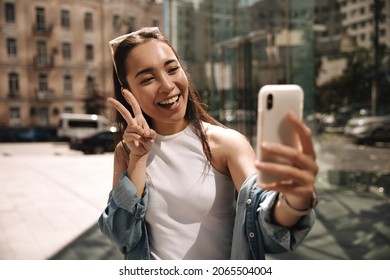 Excited Young Asian Girl In White T-shirt Using Smartphone Makes Selfie Showing Two Fingers On Street. Cute Lady Takes Pictures Of Herself And Looks At Phone Screen.