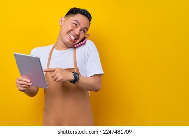 Excited young Asian man barista barman employee wearing brown apron working in coffee shop talk on mobile phone with using digital tablet to accept online orders on yellow background. business startup - Powered by Shutterstock