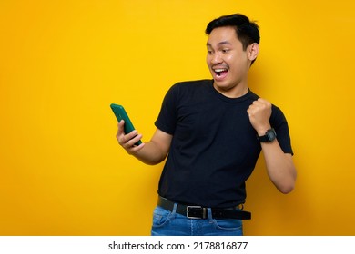 Excited Young Asian Man In Casual T-shirt Holding Mobile Phone, Making Winner Gesture, Getting Good News Isolated On Yellow Background. People Lifestyle Concept