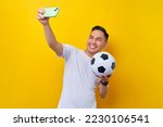 excited young Asian man football fan in a white t-shirt Carrying a soccer ball and doing a selfie shot on a mobile phone isolated on yellow background. People sport leisure lifestyle concept